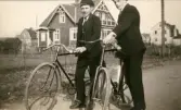 Två unga män står med sina cyklar på en grusig landsväg. Lägg märke till det stora huset i bakgrunden med snickarglädje under takåsarna. Båda männen bär huvudbonader och mörka kostymer. Den som står längst åt vänster och som bär skärmmössa, är Åke Börjesson.
Fotografiet ser ut att vara taget under slutet av 1920-talet eller 1930-talets början. 
Lägg gärna märke till cyklarnas utseende: Mycket är sig likt, men cykelstyrena och själva däcken ser annorlunda ut om man jämför med en nutida cykel. 