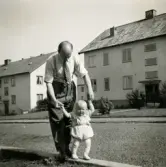 Farfar Bertil Berndtsson hjälper barnbarnet Lennart Berndtsson att gå. Bostadshus i bakgrunden. Prytzgatan i Broslätt, Mölndal, år 1954.