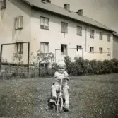 Porträtt av Lennart Berndtsson som sitter på en trehjuling framför ett bostadshus. Prytzgatan i Broslätt, Mölndal, ca 1955.