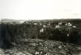 Vy över öppet landskap med bostäder, kyrka och sjö i bakgrunden samt skog och berg i förgrunden, troligen bild över Bollnäs på 1920-talet.