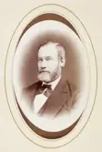 En man i helskägg, klädd i mörk kostym väst och fluga.
Bröstbild, halvprofil. Ateljéfoto.
Rektor Henrik Cederschiöld (1824-1900), Växjö.