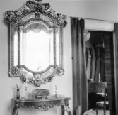 Rikt dekorerad väggspegel över ett konsollbord. Genom ett draperi bredvid möblemanget ser man in i ett herrum. Interiörbild från Torborg Lindéns hem på Södra Ringgatan 12, kv Trädgården, Alingsås. Bilden tagen av hennes son Erik Lindén 1949.