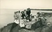 En grupp personer, både män och kvinnor, som sitter på en klippformation någonstans i någon skärgård, tyder på tidigt 1900-tal med tanke på klädseln.