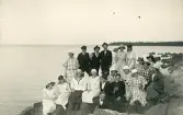 En grupp personer, både män och kvinnor, som sitter på en klippformation någonstans i någon skärgård, tyder på tidigt 1900-tal med tanke på klädseln.