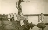 En grupp kvinnor, som sitter vid en strandkant med en träkonstruktion som går runt ett träd, tyder på tidigt 1900-tal med tanke på klädseln.