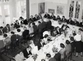 50-årsjubileum den 3 juli 1954 för Kustsanatoriet Apelviken. Middag vid långbord i Societetsrestaurangen. Serveringspersonalen serverar glas med dryck från stora brickor. Vid väggen står en skärm med mordern konst. (Se bildnr KSA137-KSA159.)