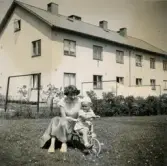 Porträtt av Lennart Berndtsson och hans mor Inga Maj Berndtsson. De sitter på en trehjuling i gräset mellan husen i ett bostadsområde. Prytzgatan i Broslätt, Mölndal, cirka 1955.