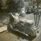 Pojken Lennart Berndtsson sitter på en motorcykel som står i en trädgård, cirka 1955. Fotografi eventuellt taget i Lödöse.