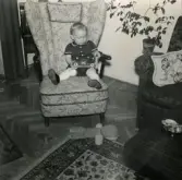 Pojken Lennart Berndtsson sitter i en fåtölj med finkläderna på i ett vardagsrum. I knät har han en leksaksbil. Tolsegårdsgatan 4 i Bosgården, Mölndal, hösten 1955.