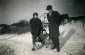 Porträtt av pojken Lennart Berndtsson tillsammans med hans mor Inga Maj Berndtsson och hans far Leif Berndtsson. Lennart sitter i en sulky. Bebyggelse i bakgrunden. Fotografi taget nära Åbybergskullen i Mölndal, en vinterdag cirka 1956.