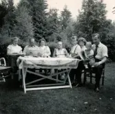 Porträtt av hela familjen samlad vid ett bord i en trädgård i Lödöse, sommaren 1956. Lennart Berndtsson sitter i pappa Leif Berndtssons knä till höger.