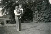 Morfar Thure Johansson står i en trädgård med barnbarnet Lennart Berndtsson i famnen. En hundkoja och en hund i bakgrunden. Oskarström cirka 1956.