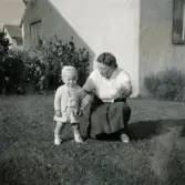 Porträtt av pojken Lennart Berndtsson och hans farmor Hildur Berndtsson i en trädgård. Bostadshus i bakgrunden. Prytzgatan i Broslätt, Mölndal, cirka 1957.