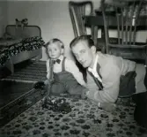 Jul på Prytzgatan 38 i Broslätt, Mölndal, cirka 1957. Pojken Lennart Berndtsson leker med en leksaksstegbil på golvet i vardagsrummet tillsammans med sin far Leif Berndtsson.