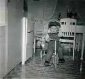 Porträtt av Lennart Berndtsson som sitter på sin trehjuling med flak i köket på Prytzgatan i Broslätt, Mölndal, julen 1957. Han är klädd i en uniform som farmor sytt samt en busschaufförsmössa. Cykeln har han fått i julklapp.