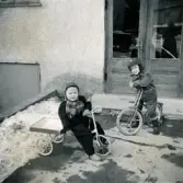 Porträtt av två pojkar som sitter på sina cyklar utanför porten till bostadshuset Prytzgatan 38 i Broslätt, Mölndal, i januari 1958. Lars Appelgren till höger. Lennart Berndtsson till vänster.