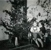 Jul på Prytzgatan i Broslätt, Mölndal, år 1957. Inga Maj Berndtsson sitter med sonen Lennart Berndtsson i knät intill julgranen i vardagsrummet och läser en bok.