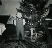 Jul på Prytzgatan i Broslätt, Mölndal, år 1957. Porträtt av den finklädde pojken Lennart Berndtsson som står intill julgranen i vardagsrummet.