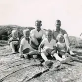Porträtt av två män och fyra barn som sitter på klipporna vid havet. Knarrholmen i Göteborgs södra skärgård, cirka 1959. Bland de sex ses pojken Lennart Berndtsson och hans far Leif Berndtsson.