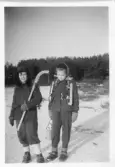 Lennart Gunnarsson och Gudrun Olsson står i ett vinterklätt landskap, troligtvis i Tutjärn, Kållered 1954. Lennart står med en bandyklubba i handen och Gudrun har ett par skridskor runt halsen. Barnen har fika med sig.