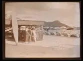 Män står en servering på en badstrand. En surfingbräda står lutad mot hyddan av flätade palmblad.