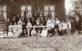 En stor grupp människor, både män, kvinnor och barn, uppställda framför en träbyggnad, kanske en stor familjesammankomst. Text baksida kort: 