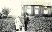 Bild av tre personer, en man, en kvinna och ett barn, som står utomhus framför en träbyggnad, troligen deras hem.