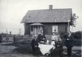 Bilden visar en grupp människor som sitter utomhus runt ett bord och fikar, bakom dem finns en stuga.