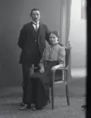 Parporträtt, ateljéfoto, beställare Alfred Hansson från Abborrakull, Ätran. Kvinnan sitter i länsstol med mannen stående bredvid.