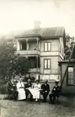 Bilden visar en grupp människor som sitter utomhus vid ett bord framför en träbyggnad med balkong och veranda, troligen början av 1900-talet.