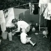 Familjen Erikssons tältsemestrar med bil på 1950- och 1960-talet. Mamma Gun-Britt håller på med hushållsbestyr vid tältet och Björn sitter brevid, i Ljugarn på Gotlands östra sida 1959.