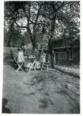 Tre personer som sitter i en lummig trädgård vid ett bord och fikar. Text baksida kort: 