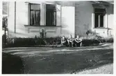 Tre personer som sitter på marken framför ett hus. Text baksida kort: Östanby 1947.
