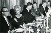 Glasdesignern Asta Strömberg med några herrar i kostym vid ett middagsbord.
På baksidan av fotot finns text: 