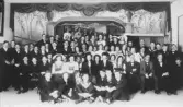 Gruppfotografi föreställande cirka 60–70 ungdomar och unga vuxna samlade på en scen i en dekorerad samlingssal. Scenen är försedd med draperad ridå och målad fond med kolonner och växtmotiv samt en lyrsymbol ovanför scenöppningen. Deltagarna är klädda i festkläder; männen i mörk kostym med fluga och kvinnorna i ljusa klänningar, några med hatt eller folkdräktsinspirerade plagg. Bilden är sannolikt tagen i samband med föreningsverksamhet, musik- eller teaterframträdande under 1910- eller 1920-tal.