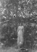 Fotografi föreställande en kvinna stående i skogsmiljö. Hon bär ljus hellång klänning med lång ärm och dekor vid halsringningen. Bilden är tagen utomhus i dagsljus med lövträd och undervegetation i bakgrunden, En hatt hänger i en gren. Sannolikt taget omkring 1910–1920 tal.