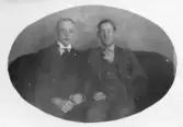 Ateljéporträtt av två män sittande bredvid varandra på stoppad soffa. Den yngre mannen bär mörk kostym med hög styv krage och slips. Den äldre mannen bär tredelad kostym med mönstrad slips. Bakgrunden utgörs av målad ateljéfond. Bilden är beskuren i oval form. Sannolikt tagen omkring 1910–1920.