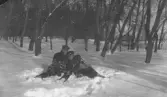 Två män sitter i djup snö i ett skogsparti vintertid. De bär mörka ytterrockar och skärmmössor. Männen sitter tätt intill varandra med armarna om varandra. Omgivningen utgörs av vissna lövträd och snötäckt mark. Bilden är tagen i naturligt ljus och har karaktär av amatörfotografi. Troligen från 1910-1920 tal.