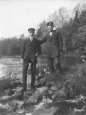 Två unga män står på stenar vid kanten av ett vattendrag. De är klädda i mörka kostymer och bär mörka skärmmössor med emblem. Den ena mannen har handen i fickan medan den andra har armen lagd över den förstes axel. I förgrunden syns rinnande vatten och stenar, och i bakgrunden träd och vegetation längs strandkanten. Bilden kan dateras till cirka 1910–1920.