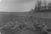 Grupporträtt av åtta uniformerade män som ligger och sitter tätt tillsammans på en gräsbevuxen mark i ett öppet landskap. De bär militäruniformer med mössor och flera av dem röker cigarett. Männen är avslappnat poserade och ser mot kameran. I bakgrunden syns öppna fält, en höjd med träd samt en byggnad, vilket tyder på att bilden är tagen utomhus i lantlig miljö. Fotografiet förefaller vara från tidigt 1900tal.