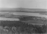 Panoramavy över ett sjö- och skogslandskap sedd från höjd. I förgrunden syns barrskog och nedanför breder flera sjöar eller älvutvidgningar ut sig med holmar och näs. På en udde ligger spridd bebyggelse bestående av gårdar och ekonomibyggnader omgivna av uppodlad mark. I bakgrunden fortsätter det kuperade landskapet med vatten och skogspartier. Fotot är taget från utsiktspunkt och dateras sannolikt till tidigt 1900-tal.
