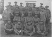 Grupporträtt av ett tjugotal uniformerade män uppställda framför en träbyggnad. Männen är klädda i militära uniformer med knäppta vapenrockar och fältmössor, och är arrangerade i flera rader – sittande på marken längst fram, sittande på bänk eller knä i mitten samt stående längst bak. Några bär gradbeteckningar på ärmar och axlar. Bilden är tagen utomhus i direkt anslutning till byggnaden, vars panel och dörr syns i bakgrunden. Fotografiet ger intryck av ett regements- eller plutonsfoto och kan dateras till tidigt 1900-tal.