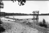 Skogaby, badplats. Badbrygga med hopptorn i sjö kantad av skog. En person går på bryggan.