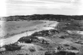 Ugglarps havsbad. Strandvy med badande och solande människor. Några småbåtar i havet och längs bergets fot ligger spridd bebyggelse. Förlag 1961: Konsum, Ugglarp.