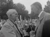 Kungabesök. Kung Gustaf VI Adolf hälsar på 90-årige f d kustroddaren Gustafson. Runt dem står barn och ungdomar med svenska flaggor. En gosse fotograferar fotografen. Artikel i samband med bilden publicerad i Varbergs Tidning 1954-06-23.