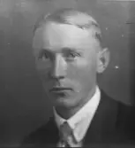 Porträtt av man i halvfigur mot neutral bakgrund. Han är klädd i mörk kavaj, ljus skjorta och slips. Blicken riktad mot kameran. Ateljéfotografi.