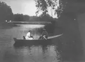 Svartvitt fotografi av tre personer och en hund i en roddbåt på lugnt vatten. En man sitter vid årorna, mittemot en kvinna i ljus klädsel och ytterligare en person. Vass och träd kantar stranden och i bakgrunden syns en bro.