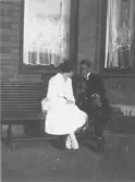 Svartvitt fotografi av en kvinna och en man sittande på en bänk utanför en tegelbyggnad med spetsgardiner i fönstren. De sitter nära och är vända mot varandra. Tidigt 1900-tal.