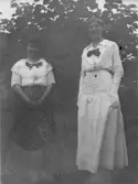 Svartvitt fotografi av två kvinnor stående utomhus framför en tät buske. Båda är klädda i ljusa blusar med stora rosetter vid kragen. Den ena bär mörk mönstrad kjol, den andra en lång ljus kjol. De står vända mot kameran. Tidigt 1900-tal.