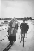 En grupp människor avbildade på skidor i snötäckt landskap. I förgrunden ses en kvinna och en man med skidstavar och vinterklädsel, gående mot kameran. I bakgrunden framträder bostadshus, stolpar och en uppbyggd träkonstruktion. Tidigt 1900-tal.